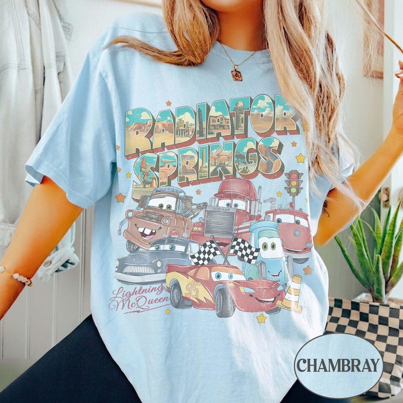 K&ouml;nnte beinhalten: Hellblaues T-Shirt mit einer Grafik von Cars-Charakteren, darunter Lightning McQueen, und dem Text "Radiator Springs". Das Shirt hat kurze &Auml;rmel und eine lockere Passform.