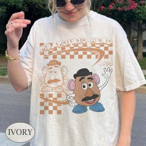 May include: An ivory t-shirt featuring a graphic print of Mr. Potato Head. The design includes a brown checkered background, the text "MR. POTATO HEAD" and "1995" in a retro font, and a colorful illustration of the character waving alongside a line-art version.