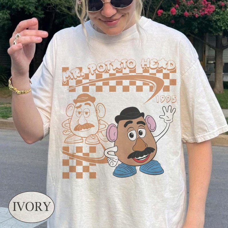 May include: An ivory t-shirt featuring a graphic print of Mr. Potato Head. The design includes a brown checkered background, the text "MR. POTATO HEAD" and "1995" in a retro font, and a colorful illustration of the character waving alongside a line-art version.