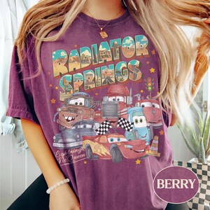 K&ouml;nnte beinhalten: Beerenfarbenes T-Shirt mit einem Aufdruck von Charakteren aus dem Film Cars, mit dem Text "Radiator Springs" und "Lightning McQueen". Das Design umfasst Autoillustrationen und Sterne.