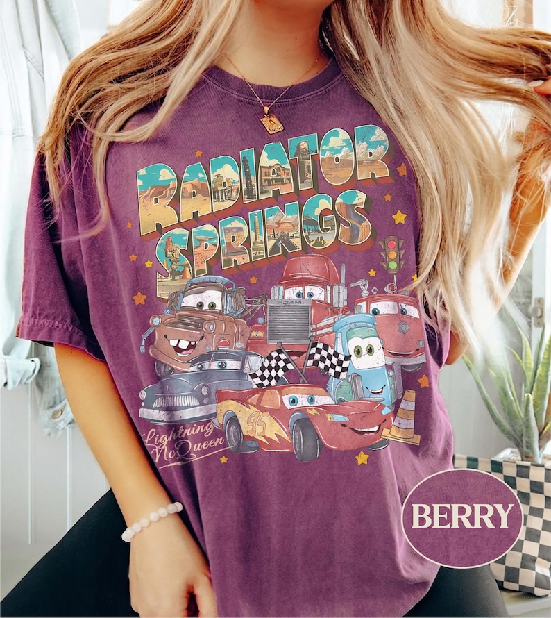 K&ouml;nnte beinhalten: Beerenfarbenes T-Shirt mit einem Aufdruck von Charakteren aus dem Film Cars, mit dem Text "Radiator Springs" und "Lightning McQueen". Das Design umfasst Autoillustrationen und Sterne.