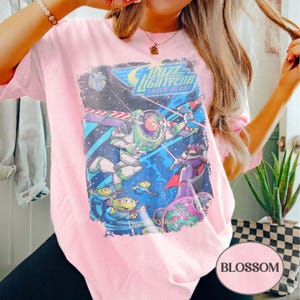 Puede incluir: Camiseta rosa claro con un estampado gr&aacute;fico de Buzz Lightyear de Toy Story. El dise&ntilde;o incluye el texto "Buzz Lightyear Laser Blast" y "Disneyland". El gr&aacute;fico est&aacute; situado sobre un fondo espacial con varios personajes.