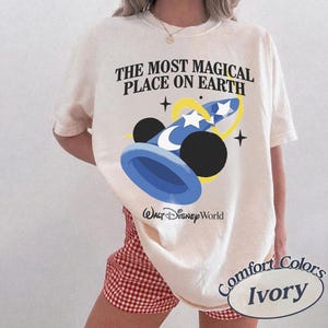 Puede incluir: Camiseta de color marfil con el texto "THE MOST MAGICAL PLACE ON EARTH" y un gr&aacute;fico azul y amarillo de un sombrero de mago con orejas de Mickey Mouse. La camiseta tambi&eacute;n tiene las palabras "Walt Disney World" y "Comfort Colors Ivory".