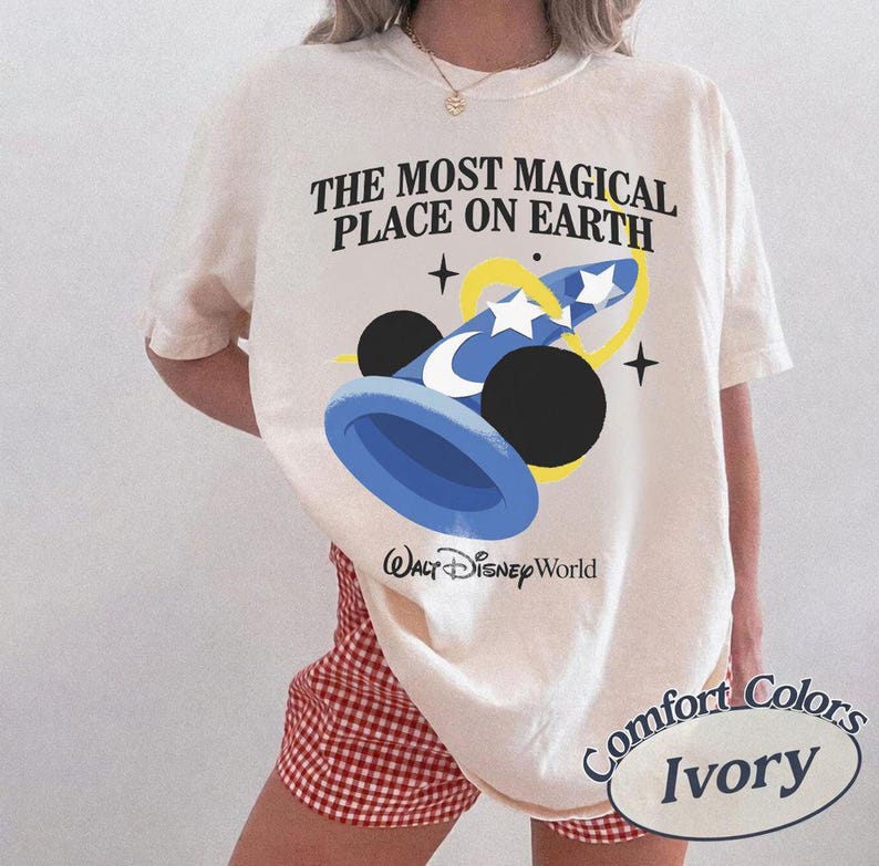 Puede incluir: Camiseta de color marfil con el texto "THE MOST MAGICAL PLACE ON EARTH" y un gr&aacute;fico azul y amarillo de un sombrero de mago con orejas de Mickey Mouse. La camiseta tambi&eacute;n tiene las palabras "Walt Disney World" y "Comfort Colors Ivory".