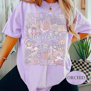K&ouml;nnte beinhalten: Orchideenfarbenes T-Shirt mit einer farbenfrohen Grafik mit Universal Studios-Charakteren und -Attraktionen. Das Design enth&auml;lt den Text "UNIVERSAL Studios". Das Shirt ist ein kurz&auml;rmeliger, l&auml;ssiger Stil.