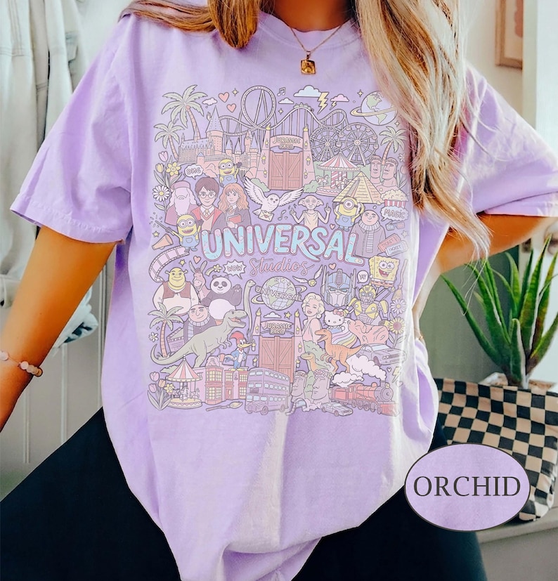 K&ouml;nnte beinhalten: Orchideenfarbenes T-Shirt mit einer farbenfrohen Grafik mit Universal Studios-Charakteren und -Attraktionen. Das Design enth&auml;lt den Text "UNIVERSAL Studios". Das Shirt ist ein kurz&auml;rmeliger, l&auml;ssiger Stil.
