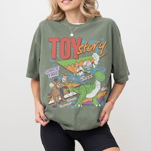 May include: Olive green t-shirt featuring the words "TOY STORY" in red and orange, with the text "SINCE 1995". The shirt has a graphic of Woody, Buzz Lightyear, and Rex the dinosaur.