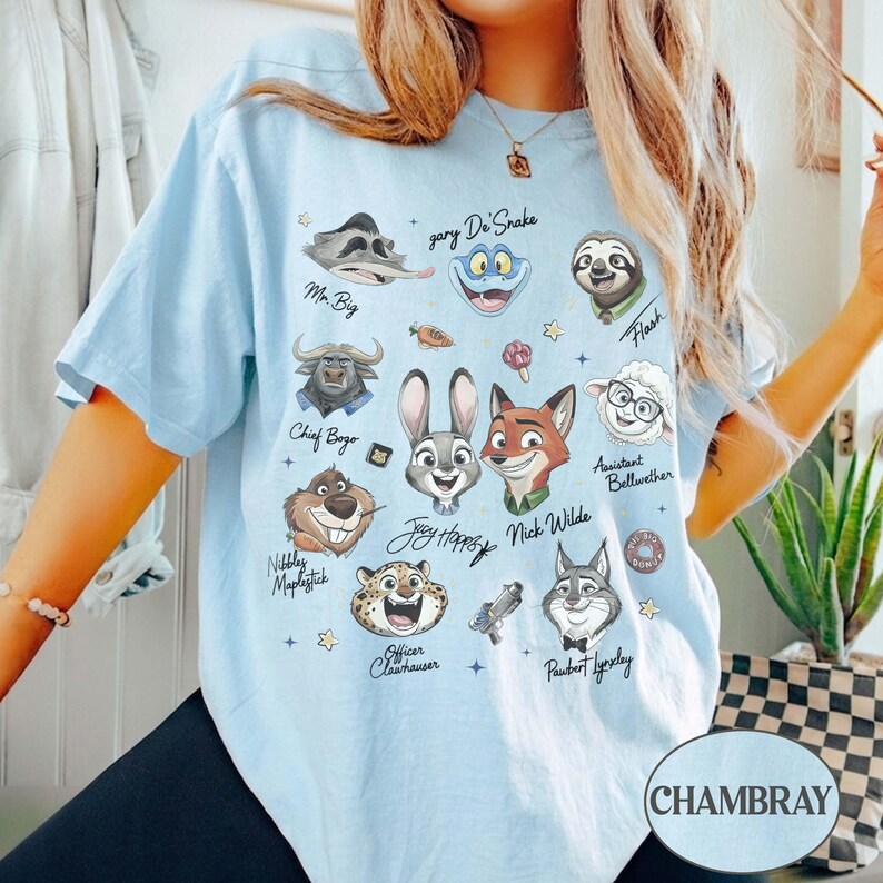 May include: A light blue t-shirt featuring a graphic print of various animated characters from Zootopia, including Judy Hopps, Nick Wilde, and Chief Bogo, with their names written in cursive script alongside small icons like a carrot and a donut.