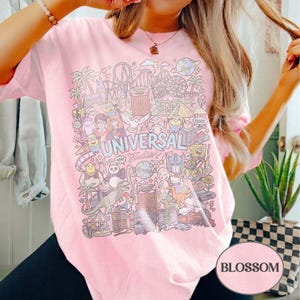 K&ouml;nnte beinhalten: Hellrosa T-Shirt mit einer farbenfrohen Illustration von Universal Studios-Charakteren und -Attraktionen. Das Design enth&auml;lt den Text "UNIVERSAL Studios" in einer verspielten Schriftart, umgeben von Cartoonfiguren und Themenparkelementen.