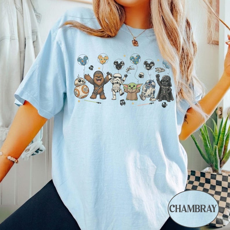May include: Light blue t-shirt featuring a Star Wars themed graphic. The graphic includes cartoon versions of characters like BB-8, Chewbacca, and Darth Vader, holding balloons. The word "CHAMBRAY" is printed on the bottom right of the shirt.