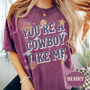 May include: A berry-colored short-sleeve t-shirt featuring the text "YOU'RE A COWBOY LIKE ME" in a bold font. The graphic includes illustrations of cowboy boots, a ball, a sheriff star, clouds, and a rocket ship, all in a vintage-inspired style.