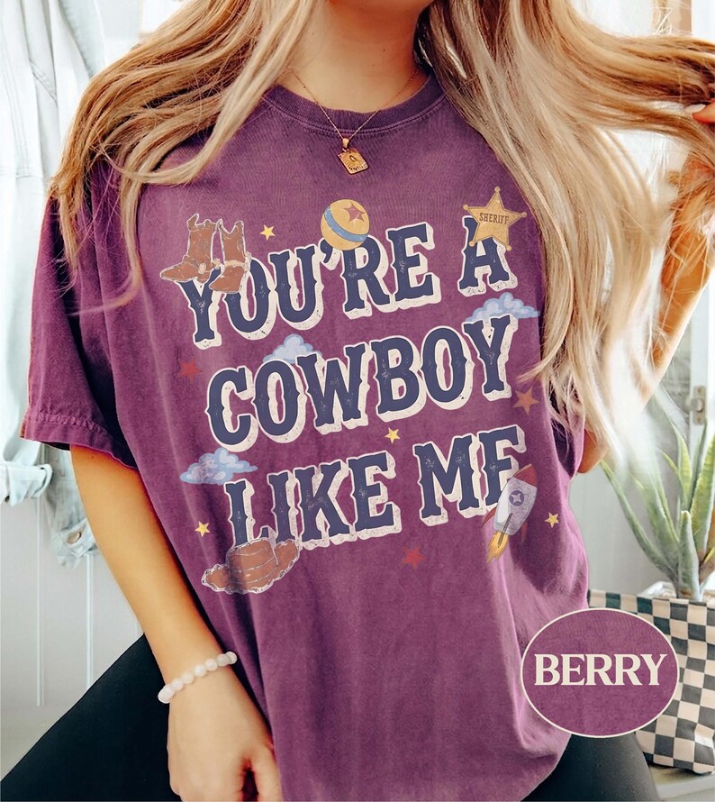 May include: A berry-colored short-sleeve t-shirt featuring the text "YOU'RE A COWBOY LIKE ME" in a bold font. The graphic includes illustrations of cowboy boots, a ball, a sheriff star, clouds, and a rocket ship, all in a vintage-inspired style.