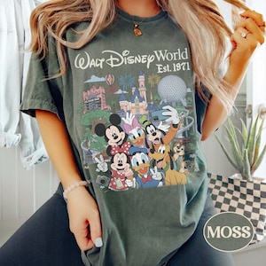 May include: A dark green t-shirt featuring a graphic print of Walt Disney World Est. 1971 with various cartoon characters including Mickey Mouse, Minnie Mouse, Donald Duck, Daisy Duck, Goofy, Pluto, and Chip and Dale taking a group selfie.