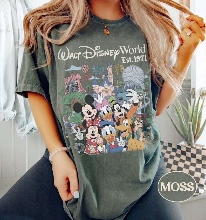 May include: A dark green t-shirt featuring a graphic print of Walt Disney World Est. 1971 with various cartoon characters including Mickey Mouse, Minnie Mouse, Donald Duck, Daisy Duck, Goofy, Pluto, and Chip and Dale taking a group selfie.