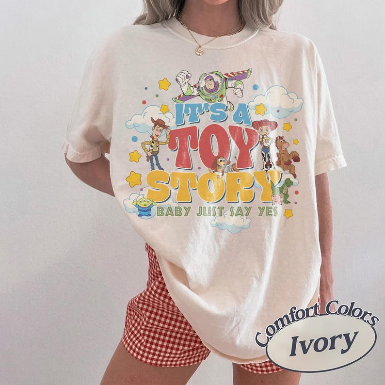 Peut inclure: Un t-shirt ivoire avec une impression graphique color&eacute;e de personnages de Toy Story, dont Buzz l'&Eacute;clair, Woody, Jessie et un alien. Le texte indique IT'S A TOY STORY BABY JUST SAY YES. Le t-shirt est port&eacute; avec un short &agrave; carreaux vichy rouge et blanc.