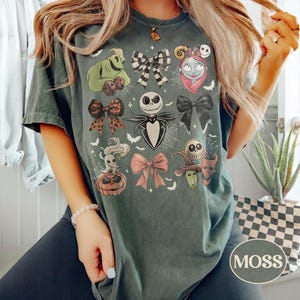 May include: A dark green t-shirt featuring a graphic print of characters and bows from The Nightmare Before Christmas. The design includes Jack Skellington, Sally, Oogie Boogie, Zero, and various bows arranged in a grid pattern with spiderwebs.