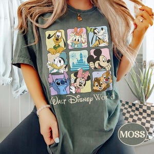 May include: Moss green t-shirt featuring a grid of Disney characters including Mickey Mouse, Donald Duck, and Stitch. The words "Walt Disney World" are printed below the grid. The shirt has a crew neck and short sleeves.