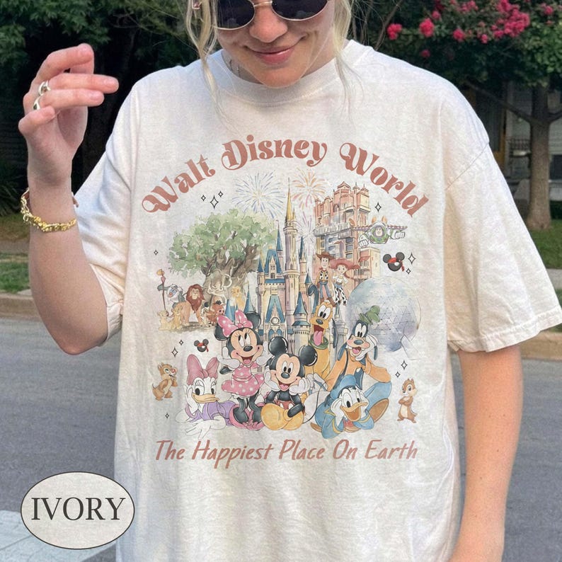 May include: Ivory-colored t-shirt featuring a colorful graphic of Disney characters and the text "Walt Disney World" and "The Happiest Place On Earth." The design includes Mickey and Minnie Mouse, Donald Duck, Goofy, and other characters.