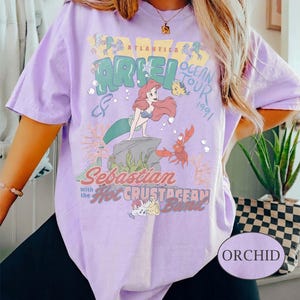 May include: A light purple t-shirt featuring a graphic print of Ariel, Flounder, and Sebastian. The text reads Atlantica Ocean Tour 1991, Ariel, and Sebastian with the Hot Crustacean Band. The design includes coral, bubbles, and sea elements in muted tones.