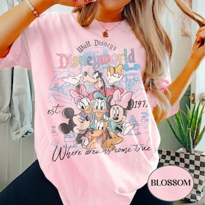 May include: A light pink t-shirt featuring a colorful graphic of Disney characters, including Mickey Mouse, Minnie Mouse, and Goofy. The text "Walt Disney's Disneyworld" and "Where dreams come true" are also displayed. The shirt is a crew neck.