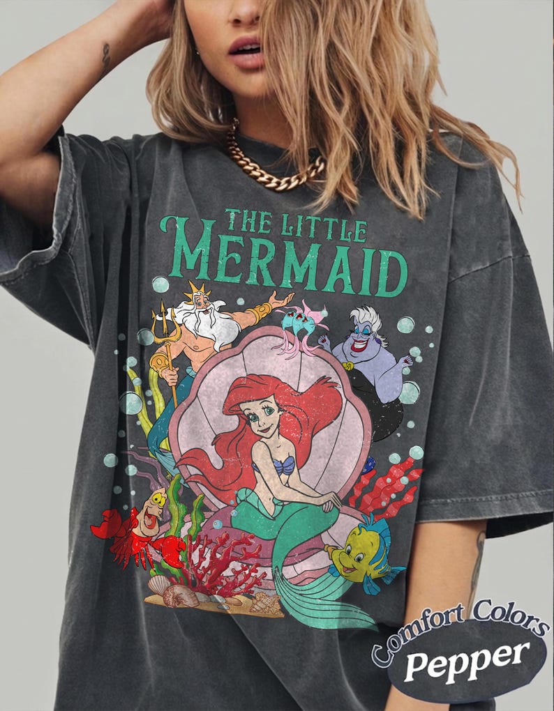May include: Dark gray t-shirt featuring a graphic of Ariel and other characters from Disney's The Little Mermaid. The design includes the text "THE LITTLE MERMAID" in green. The shirt is a Comfort Colors Pepper tee.
