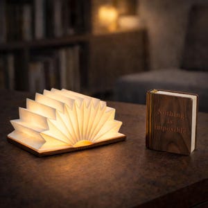 May include: A wooden book-shaped lamp with open, illuminated pages, resembling a fan. Beside it, a closed book with the words "Nothing is impossible" is displayed. Both items are on a dark brown surface, creating a warm, inviting ambiance.