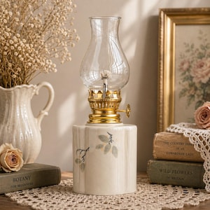 May include: A vintage-style oil lamp with a clear glass globe and a gold-colored metal base. The cream-colored ceramic base features a floral design. The lamp is displayed with a vase of dried flowers and antique books.