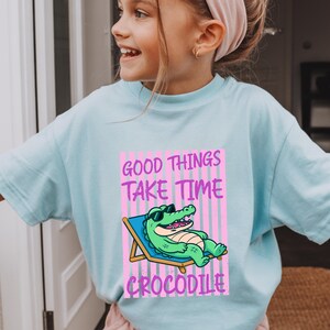 May include: A light blue t-shirt featuring a graphic print of a green crocodile wearing sunglasses while lounging on a blue beach chair. The background of the graphic consists of vertical pink stripes, and the text reads Good Things Take Time Crocodile.
