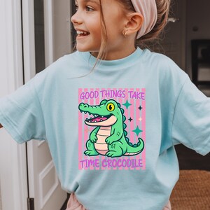 May include: A light blue t-shirt featuring a graphic of a green crocodile sitting in front of pink vertical stripes. The text above the crocodile reads Good Things Take and the text below reads Time Crocodile, accompanied by small star icons.