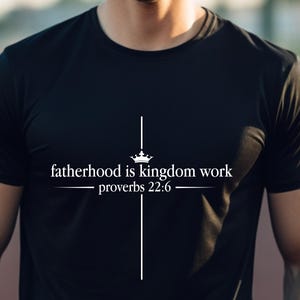 Könnte beinhalten: Ein schwarzes Kurzarm-T-Shirt mit einem weißen Grafikdesign, das ein Kreuz mit einer Krone an der Spitze zeigt. Der Text "fatherhood is kingdom work" und "proverbs 22:6" ist in einer weißen Serifenschrift horizontal über dem Kreuz zentriert.