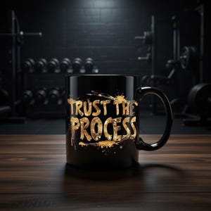 Op de afbeelding: Zwarte koffiemok met de gouden tekst "TRUST THE PROCESS". De mok heeft een zwart oor en staat tegen een donkere achtergrond met fitnessapparatuur. De tekst heeft een spatontwerp.