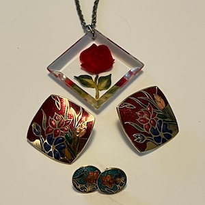 May include: A jewelry set featuring a necklace with a red rose pendant, two pairs of earrings. The earrings and pendant have a floral design with red, blue, and gold accents. The necklace chain is silver.
