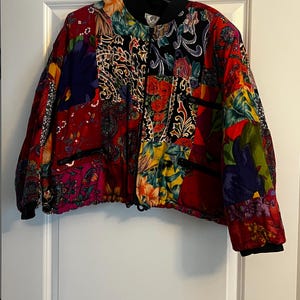 May include: A quilted bomber jacket featuring a vibrant patchwork design with various floral and paisley patterns in shades of red, yellow, blue, and black. The jacket has a black ribbed collar, black cuffs, and two zippered pockets on the front.