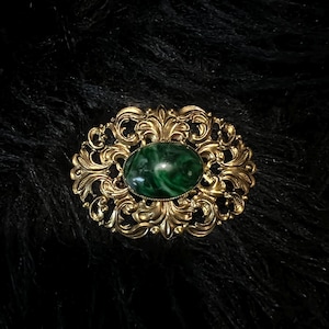 May include: An oval brooch featuring a central dark green marbled stone set within an ornate gold-toned metal frame. The metalwork displays intricate swirling scroll patterns and filigree details surrounding the smooth, polished green cabochon.