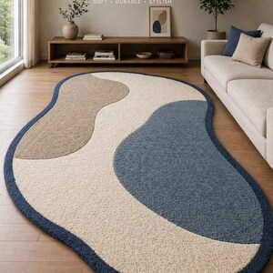 May include: Hand-tufted rug with a modern design. The rug features a unique, organic shape with sections of beige, off-white, and blue. The rug is bordered with a dark blue trim. The text "SOFT • DURABLE • STYLISH" is visible.
