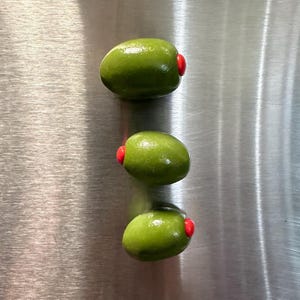 Può includere: Tre olive verdi con peperoni rossi sono disposte verticalmente su una superficie metallica spazzolata. Le olive sono di forma ovale e hanno un aspetto lucido. I peperoni rossi offrono un contrasto di colore.