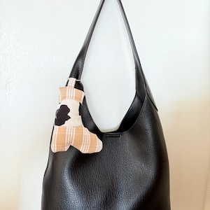 May include: Black leather shoulder bag with a single strap. A small, decorative charm with a cow print and tan plaid pattern hangs from the strap. The bag is hanging against a white wall.