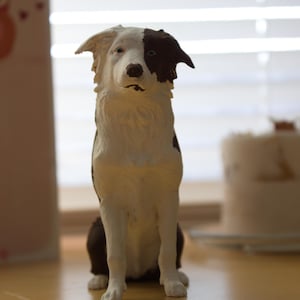 May include: A ceramic dog figurine in a sitting position. The dog has a white body with a brown patch over one eye and ear. The figurine is on a wooden surface, with a window and blinds in the background.