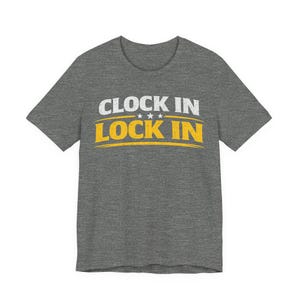 May include: A grey t-shirt featuring the text "CLOCK IN LOCK IN" in white and yellow. The text is centered on the front. The shirt has a crew neck and short sleeves. The fabric is a heathered blend.