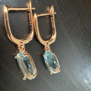 May include: A pair of rose gold earrings with a hinged closure. Each earring features a light blue, oval-shaped gemstone pendant. The earrings are displayed against a dark background.