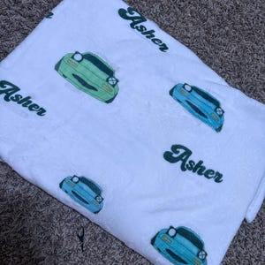 May include: White blanket with a repeating pattern of cartoon cars in shades of green and blue. The name "Asher" is printed in green script across the blanket. The blanket is made of a soft, plush material.