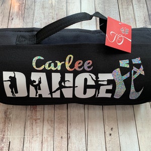 May include: Black dance bag with a white inscription that reads "Carlee DANCE" with a silhouette of dancers. The bag has a black handle and a sparkly silver dance shoe design.