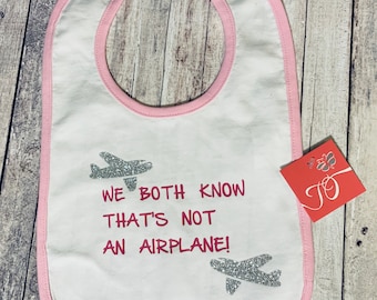 May include: White baby bib with pink trim and a silver glitter airplane design. The bib has the text "We both know that's not an airplane!" printed on it.