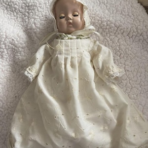 Op de afbeelding: Een vintage babypop met een cr&egrave;mekleurige jurk en muts. De jurk heeft delicate bloemenborduurwerk en oogjesdetails. Het gezicht van de pop heeft gesloten ogen en een serene uitdrukking, met een zachte, geschilderde teint. De muts is afgezet met kant.