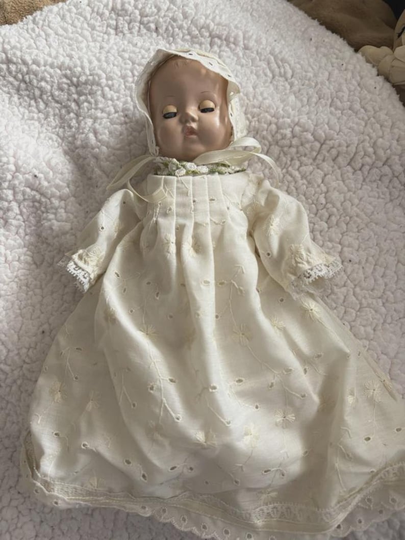 Op de afbeelding: Een vintage babypop met een cr&egrave;mekleurige jurk en muts. De jurk heeft delicate bloemenborduurwerk en oogjesdetails. Het gezicht van de pop heeft gesloten ogen en een serene uitdrukking, met een zachte, geschilderde teint. De muts is afgezet met kant.