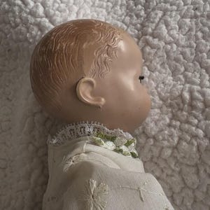 Op de afbeelding: Een vintage poppenhoofd, gezien in profiel, met een geschilderd gezicht en gevormd haar. De pop draagt een witte jurk met geborduurde bloemendetails en een kanten kraag met groene accenten. De huid van de pop is een licht perzikkleur.