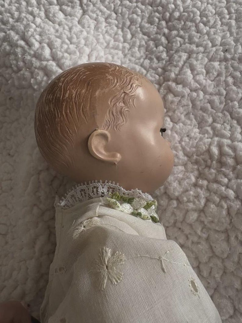 Op de afbeelding: Een vintage poppenhoofd, gezien in profiel, met een geschilderd gezicht en gevormd haar. De pop draagt een witte jurk met geborduurde bloemendetails en een kanten kraag met groene accenten. De huid van de pop is een licht perzikkleur.