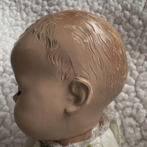 Op de afbeelding: Een vintage poppenhoofd, gezien in profiel, met een lichte huidskleur en gedetailleerde gelaatstrekken. De pop heeft gevormd haar en draagt een witte jurk met bloemenborduurwerk rond de kraag. Het poppenhoofd is ongeveer 10 cm hoog.