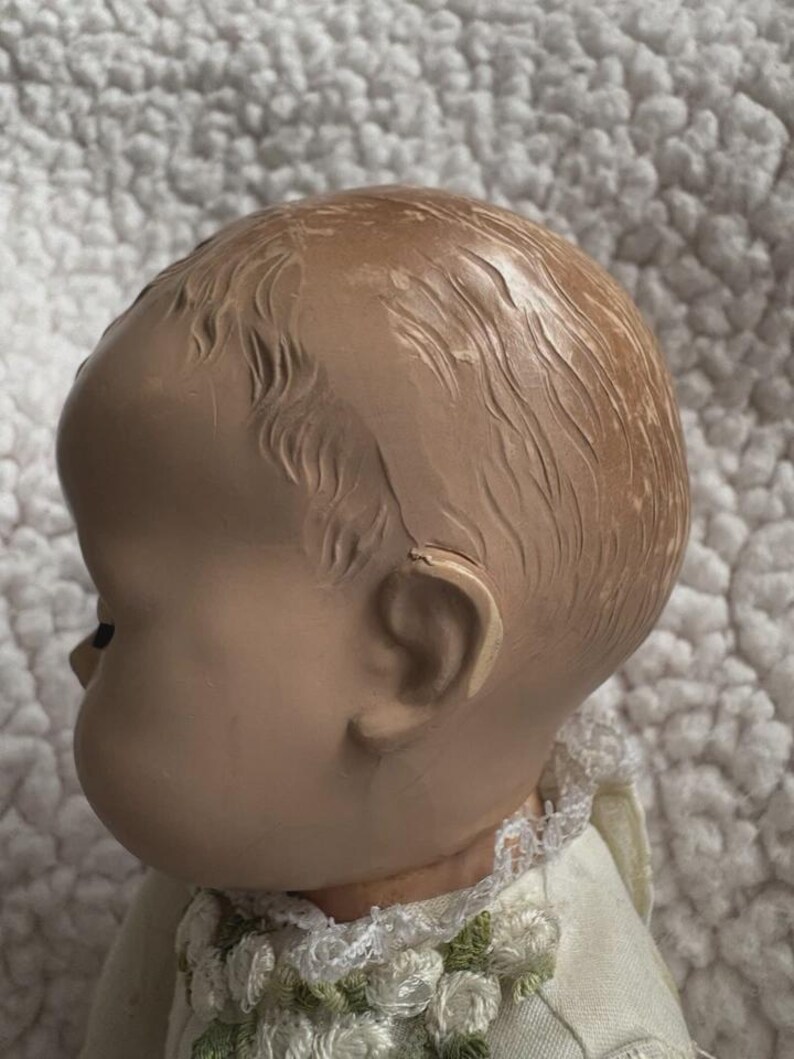 Op de afbeelding: Een vintage poppenhoofd, gezien in profiel, met een lichte huidskleur en gedetailleerde gelaatstrekken. De pop heeft gevormd haar en draagt een witte jurk met bloemenborduurwerk rond de kraag. Het poppenhoofd is ongeveer 10 cm hoog.