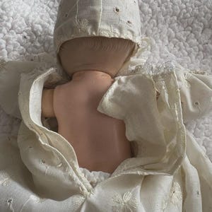 Op de afbeelding: Een vintage pop met een cr&egrave;mekleurige jurk en muts. De jurk en muts van de pop hebben delicate geborduurde bloempatronen en uitgesneden details. De huid van de pop is licht perzik van kleur.